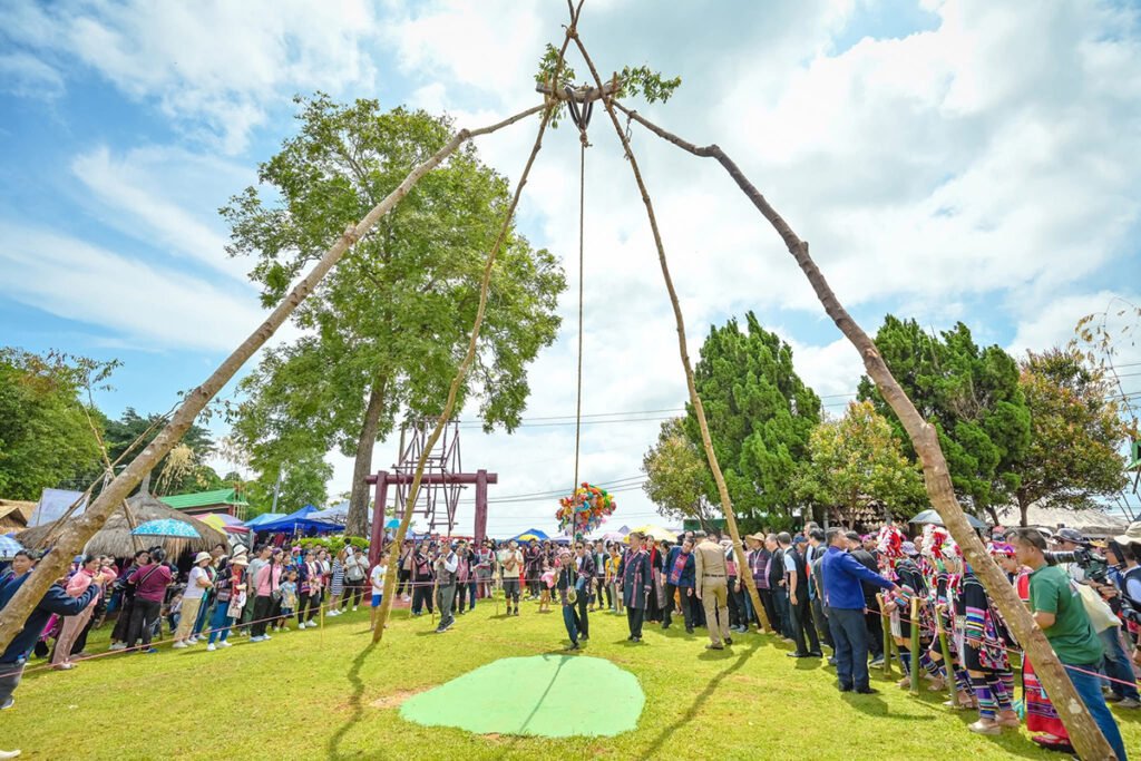 Akha villagers gather around a tall wooden swing structure during the festival, with clear skies and green trees in the background.