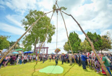 Akha villagers gather around a tall wooden swing structure during the festival, with clear skies and green trees in the background.