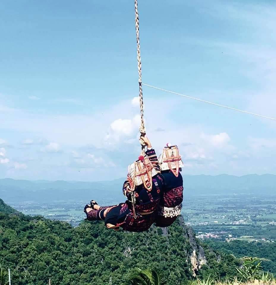 Two Akha women in traditional dress swing together above a scenic mountain landscape.