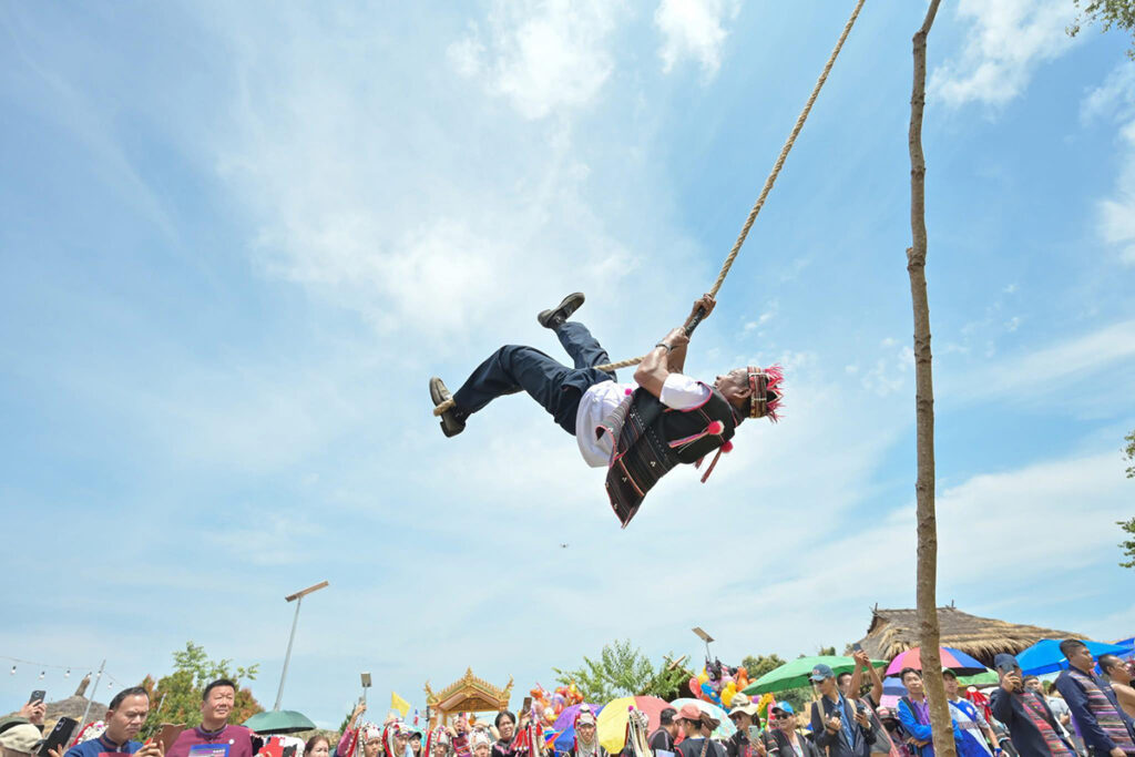 An Akha man swings high into the air on a rope swing, surrounded by festival-goers watching from below.