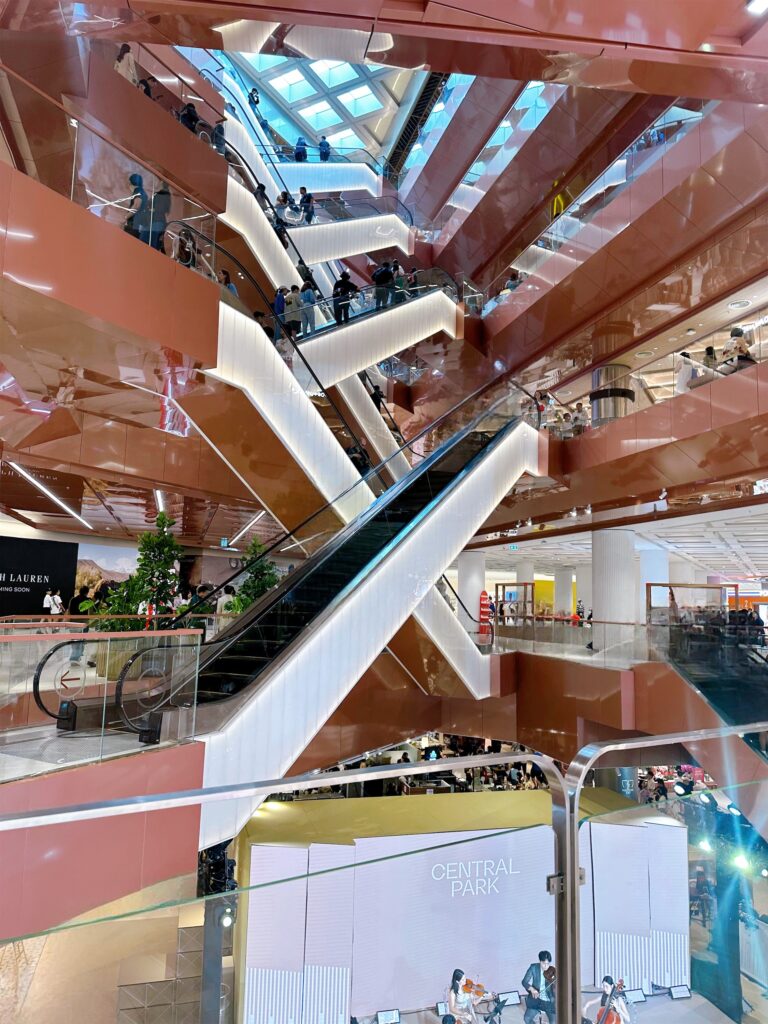 Shoppers on escalators inside Central Park Bangkok with event stage below.
