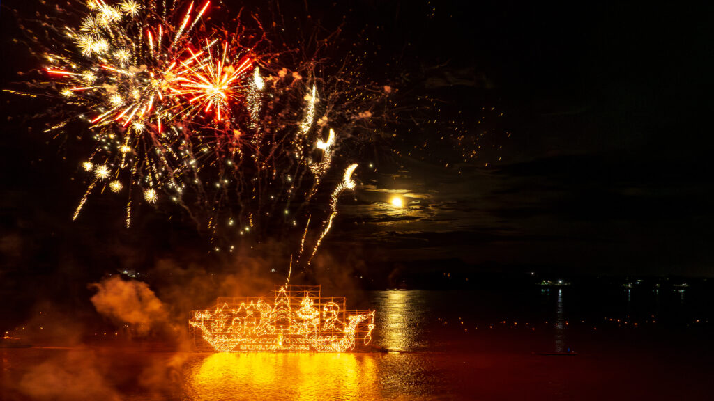 Illuminated boat parade glowing on the Mekong River with fireworks during Nakhon Phanom festival night.