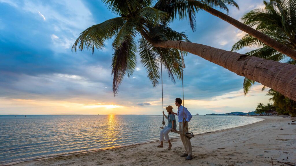 Couple relaxing on a swing beneath palm trees at Na Thon Beach, Ko Samui, during a romantic tropical sunset.