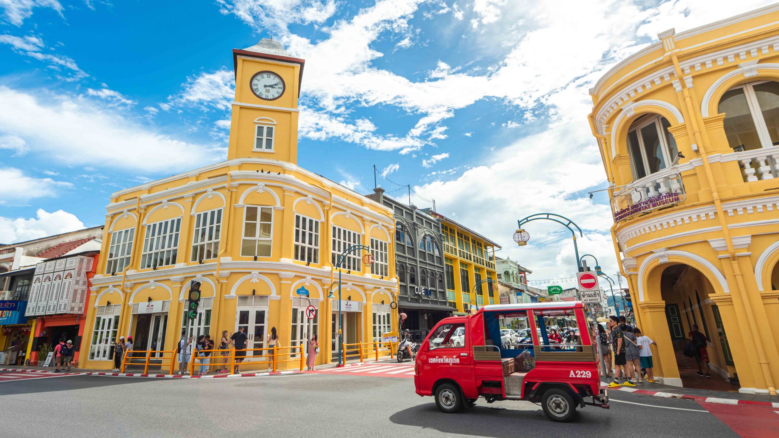 Vibrant yellow Sino-Portuguese buildings and red songthaew in Phuket Old Town under a bright blue sky.