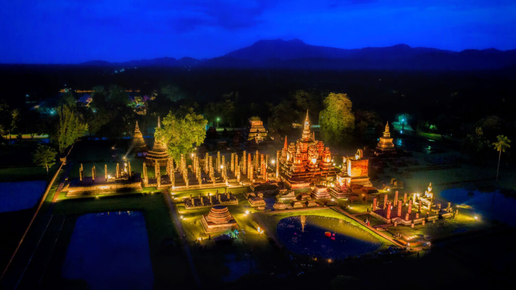 The ancient ruins of Sukhothai illuminated with golden lights against a deep blue evening sky.