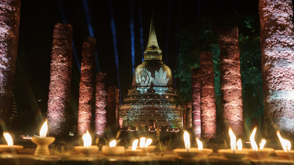 Clay candles glowing before a stupa projection at Sukhothai Historical Park during a solemn ceremony.