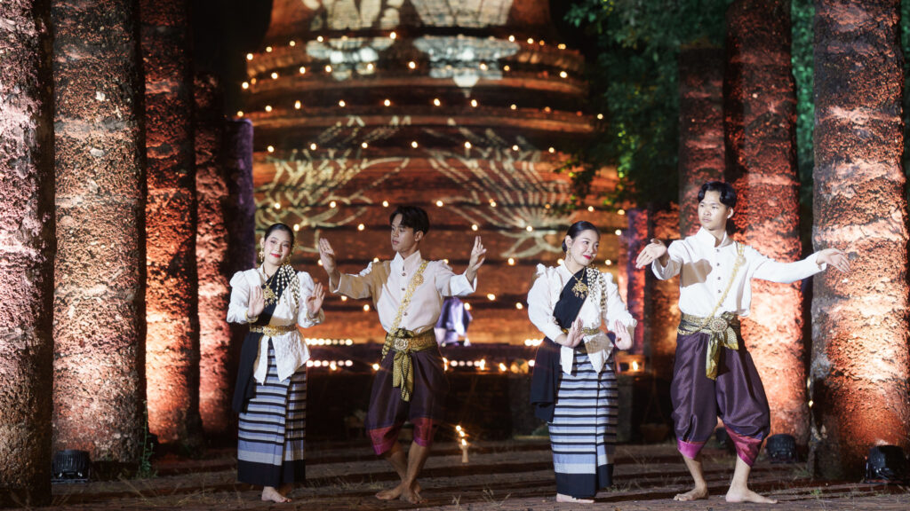 Performers in traditional attire gracefully dance amid illuminated ancient pillars at Sukhothai Historical Park.