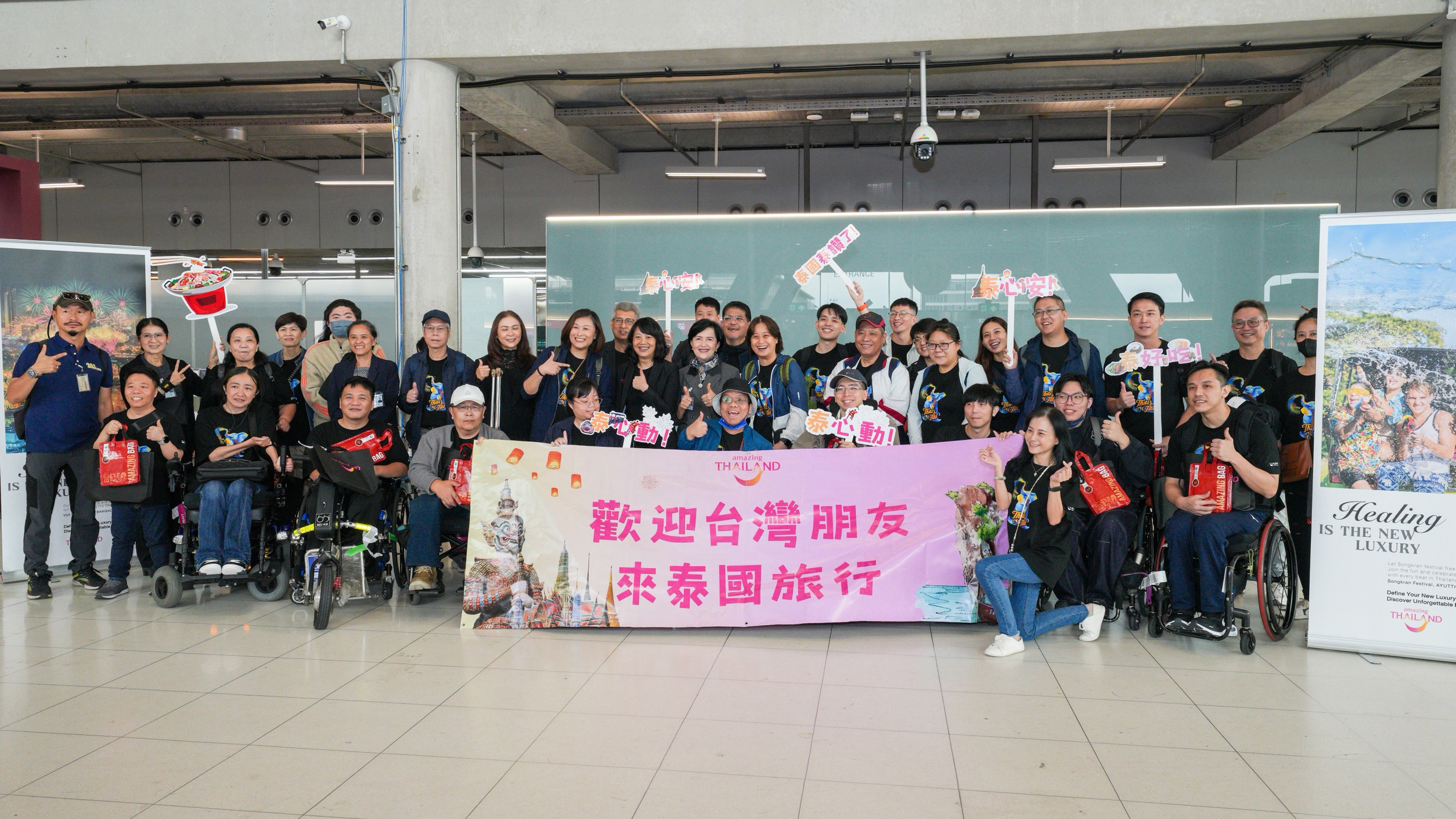 A large Taiwanese accessible tourism group poses with TAT representatives at Suvarnabhumi Airport, holding a welcome banner and “Amazing Thailand” signs.