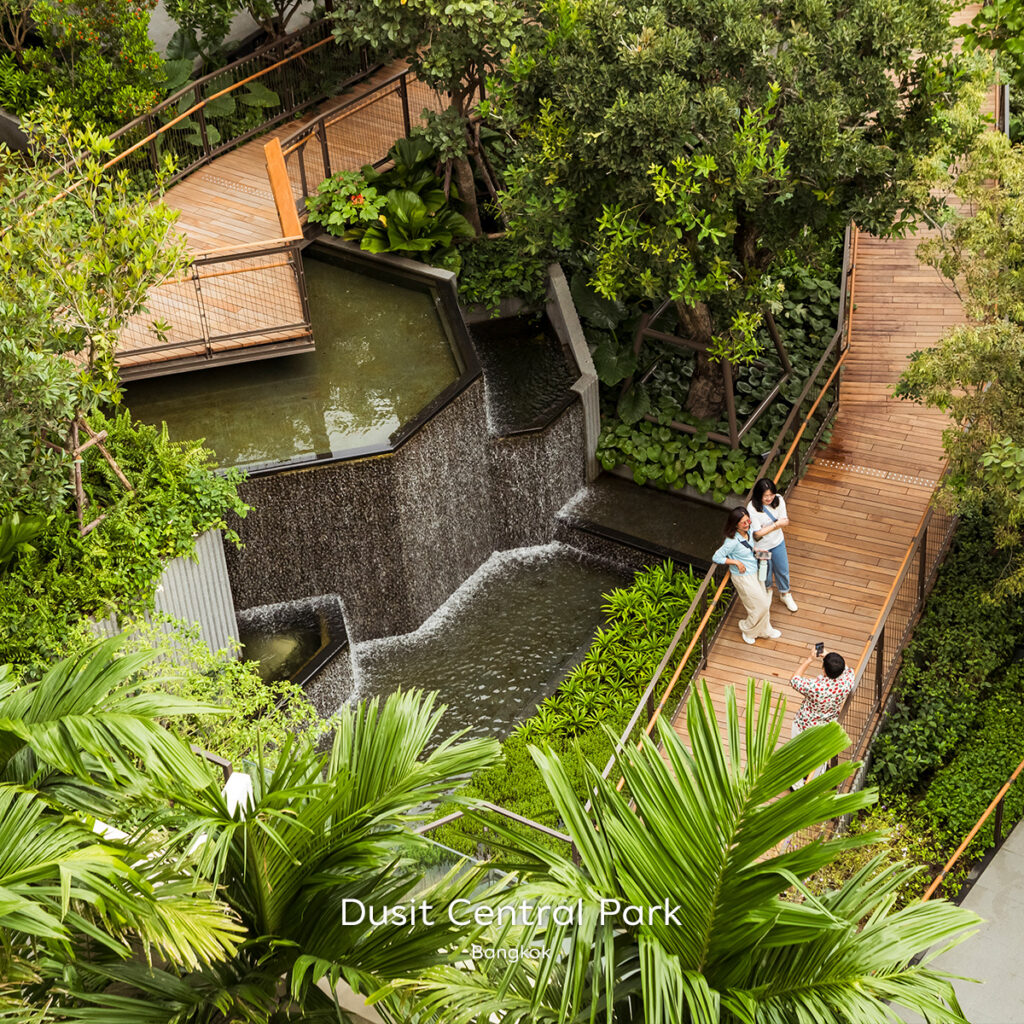 Elevated wooden walkways and tiered urban waterfall surrounded by lush greenery at Dusit Central Park in Bangkok.