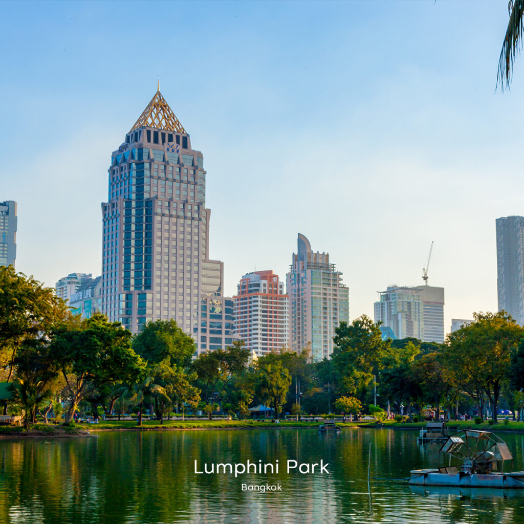 Lumphini Park lake at sunrise with lush trees and Bangkok’s skyline rising behind the water.