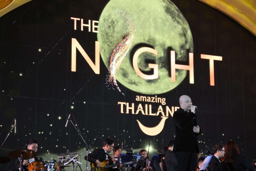 A live concert performance takes place on the main stage of The Night by Amazing Thailand, featuring a vocalist accompanied by an orchestra under moon-themed visuals.