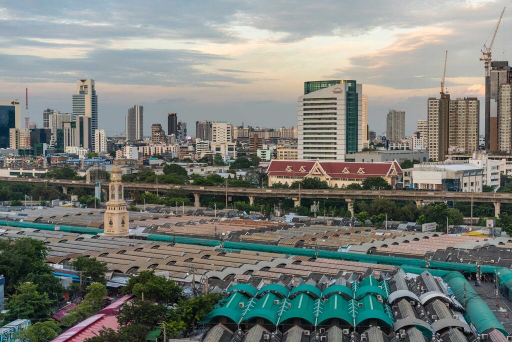 Aerial view of Chatuchak Weekend Market in Bangkok with rows of market roofs set against the city skyline at dusk.