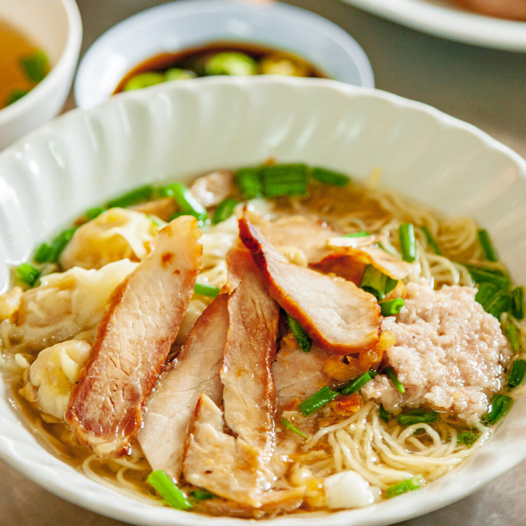 Guay Tiew rice noodle soup with roasted pork and dumplings in clear broth, highlighting Chinese influence on Thai cuisine and street food heritage.