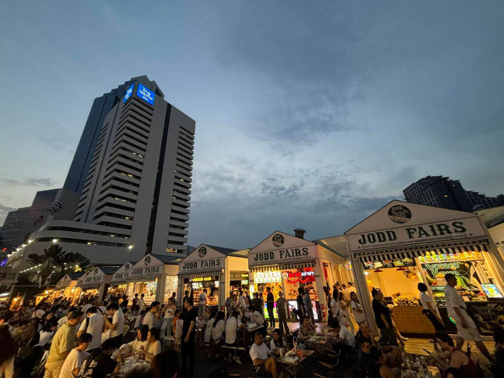 Evening atmosphere at Jodd Fairs Night Market in Bangkok with busy food stalls and diners beneath illuminated shopfronts.