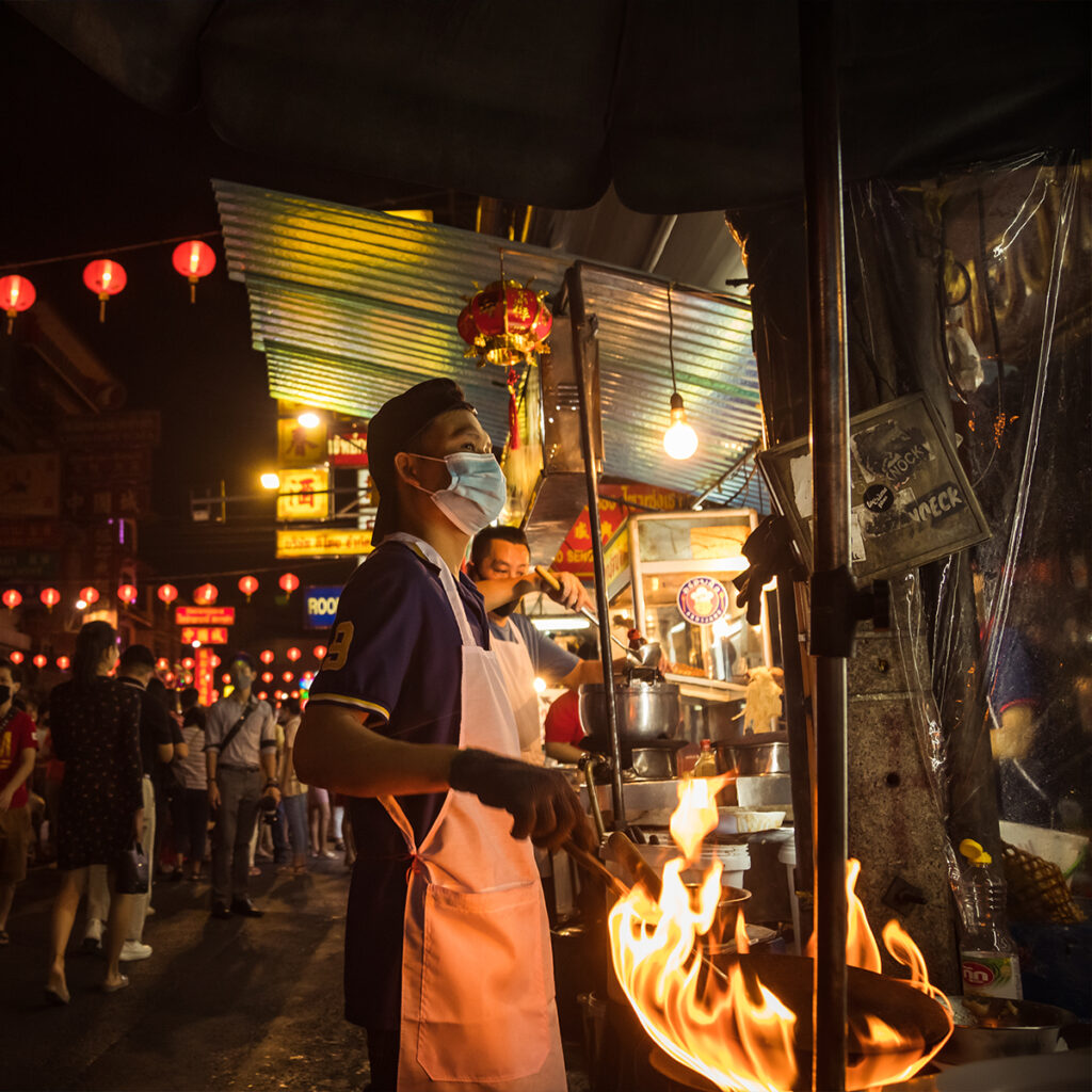 Night street food scene in Yaowarat, Bangkok, with red lanterns and wok flames showcasing Chinese influence on Thai cuisine.