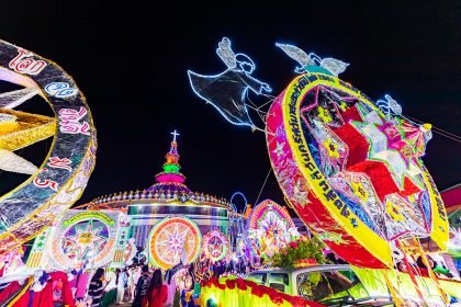 Illuminated star floats parade past the Cathedral of St Michael the Archangel during the Sakon Nakhon Christmas Star Parade, with angel motifs and vibrant lights lighting up the night sky.